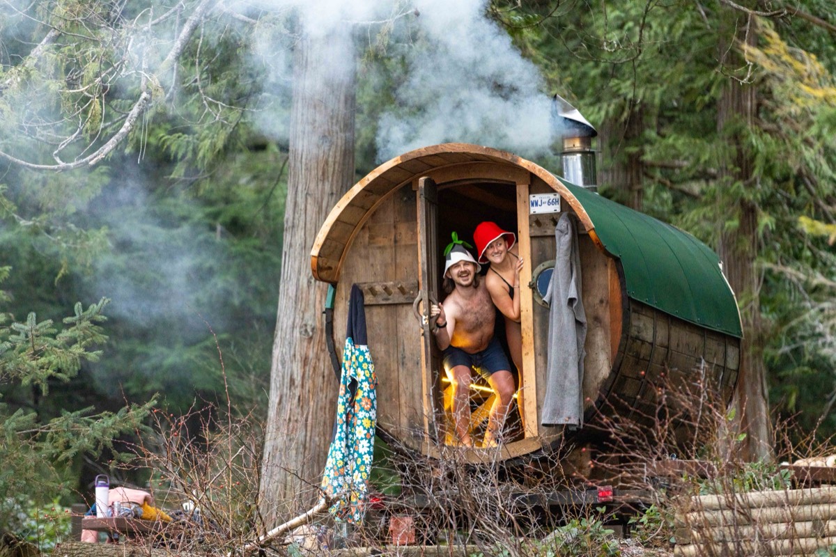 Two people in sauna hats smiling from the doorway of the wood-fired barrel sauna, steam rising