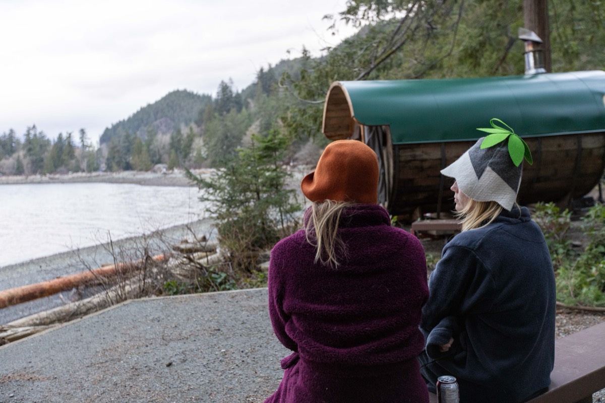 Two friends in felt sauna hats cooling down beside the barrel sauna on the shore
